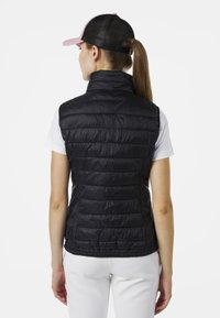 Schwarze, gepolsterte Weste mit hohem Kragen, die eine horizontale Steppung aufweist. Leichtes Material, taillierte Passform, kombiniert mit einem weißen Hemd darunter.