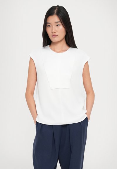Woman with long dark hair wearing a white sleeveless top and navy blue pleated trousers, standing with hands in pockets on white background.
