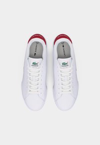 Hvite Lacoste-sneakers med hvite lisser, røde hælfaner og grønn krokodille-logo på tungen, sett ovenfra på ensfarget bakgrunn.