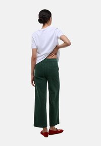 Pantalon large en velours côtelé vert avec une finition texturée, associé à un t-shirt blanc et des chaussures rouges à enfiler, montrant une coupe décontractée de dos.