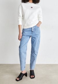 Persona vestida con una sudadera blanca de manga larga, jeans de tiro alto en un tono azul claro y mules negros de tacón abierto, de pie frente a un fondo liso.
