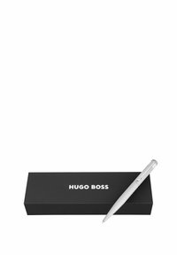 Stylo à bille en métal argenté avec une prise texturée, reposant sur une boîte noire arborant le logo blanc "HUGO BOSS". Design simple et épuré.