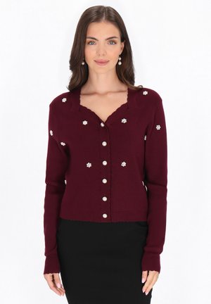 Vrouw draagt een bordeaux cardigan met parelknopen en parel bloemenversieringen, gecombineerd met een zwarte rok, staand tegen een witte achtergrond.