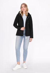 Črna zip-up jakna s kapuco, ki ima teksturirano zunanjost. V kombinaciji z svetlo modrimi kavbojkami in belimi supergami. Pod jakno je črtasta majica.