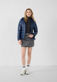 Giacca a vento blu con cappuccio, caratterizzata da un design trapuntato. Abbinata a un dolcevita nero e a una gonna in denim grigia, e a sneaker grigie con strisce nere.