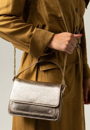 Bolso metálico dorado con acabado texturizado, solapa superior y correa de cadena. El bolso está estilizado sobre un fondo de tela marrón.