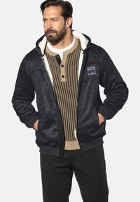 Black zip-up hoodie with a fuzzy interior, featuring a front pocket, and a tan, ribbed henley shirt underneath with buttons.