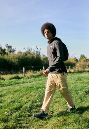 Black, hooded jacket, beige cargo pants with pockets, and black hiking shoes. Walking on grass in daylight, with a backdrop of foliage.
