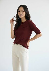 Bordowy sweter z krótkim rękawem, wykonany na drutach, ozdobiony trzema teksturowanymi sercami. W połączeniu z białymi spodniami, model trzyma okulary przeciwsłoneczne.