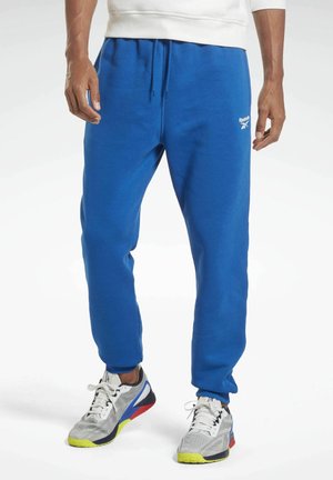 Man in blauwe Reebok joggingbroek en witte sportschoenen met meerkleurige zolen, staand met handen ontspannen langs het lichaam.
