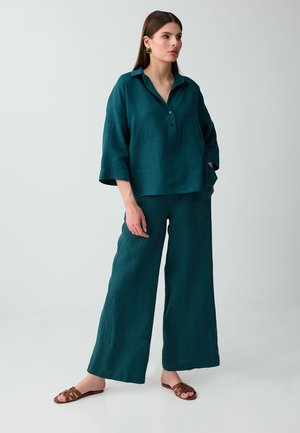 Vrouw staat en draagt een losse blauwe (teal) blouse met knopen en driekwartsmouwen en een wijde broek met bruine sandalen met open teen op een effen achtergrond.