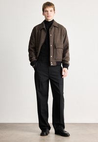 Casaco castanho texturizado com colarinho, bolso frontal e botões visíveis, combinado com calças pretas e sapatos pretos. Design simples e moderno.