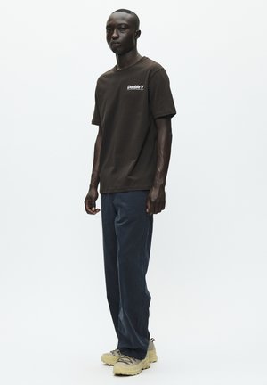 T-shirt en coton marron avec le logo "Double V" blanc, associé à un pantalon bleu foncé et des baskets chunky beige clair. Design simple, coupe confortable.