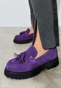 Violetti suede-loaferit fringedetaililla, joissa on paksu musta kumipohja ja teksturoitu pinta. Yhdistetty harmaisiin collegehousuihin.