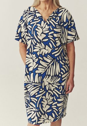 Woman wearing a blue dress with large cream tropical leaf patterns and short sleeves, standing against a plain light background.