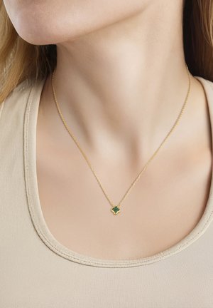 Primer plano de una persona que lleva un delicado collar de cadena de oro con un pequeño colgante de trébol de cuatro hojas verde, sobre una camiseta beige.