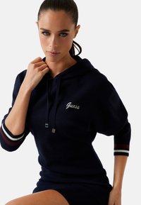 Rochie cu glugă de culoare bleumarin, cu logo-ul "Guess" brodat în alb. Prezintă accente în dungi roșii și albe pe mâneci. Fabricată dintr-un material tricotat texturat.