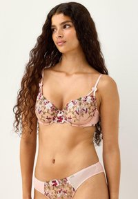 Kvinde med langt, krøllet hår iført et matchende sæt af lyserøde, blomsterbroderede bh og trusser, der kigger til venstre mod en ensfarvet baggrund.