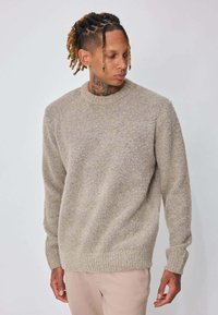Homme aux dreadlocks et tatouage au cou portant un pull beige texturé et un pantalon clair, regardant vers le bas et sur le côté sur un fond uni.