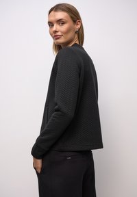 Zwarte doorgestikte jas met visgraatstructuur, ronde halslijn en lange mouwen. Heeft zijzakken en een ontwerp met losse pasvorm.