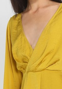 Haut enroulé jaune moutarde avec un décolleté en V profond, des manches longues et un tissu doux et texturé. Comprend des plis et des fronces à la taille.