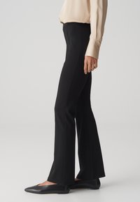 Pantalons noirs évasés en matière extensible, avec une taille ajustée et un détail de fente sur le côté. Associés à des ballerines noires élégantes.