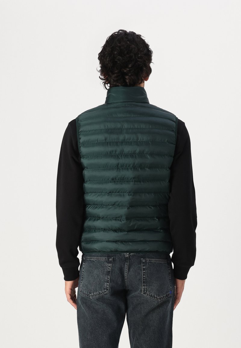 Zielona kamizelka puchowa z błyszczącego, pikowanego materiału z wysokim kołnierzem. Połączona z czarną bluzką z długim rękawem i odbarwionymi dżinsami.