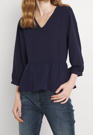 Femme portant un chemisier bleu marine à col en V avec manches 3/4 et bas à basque, associé à un jean bleu moyen, main posée sur la hanche.