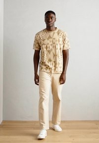 Beige Kurzarm-T-Shirt mit geometrischen Mustern, kombiniert mit hellbeigen Hosen und weißen Turnschuhen. Holzboden und neutraler Hintergrund.