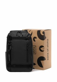 Sac à dos noir avec une finition texturée, plusieurs poches zippées, du tissu webbing à l'avant et un clip. Boîte en carton avec des motifs abstraits noirs.