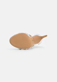 Sandale à talon haut argentée présentée de dessous, montrant une semelle beige à la texture fine et un texte de marque visible près de la voûte plantaire.
