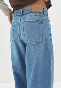 Hellblaue Jeans mit hoher Taille, weitem Bein, zwei Gesäßtaschen und einem braunen Markenpatch am Bund.