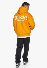 Appelsinfarvet oversized hoodie med hvid tekst "DROPSIZE CRIME CLUB" på ryggen, parret med sorte jeans og sort-hvide sneakers.