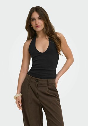 Vrouw met lang bruin haar die een zwart haltertopje en hoge bruine broek draagt, poserend met één hand op haar heup.