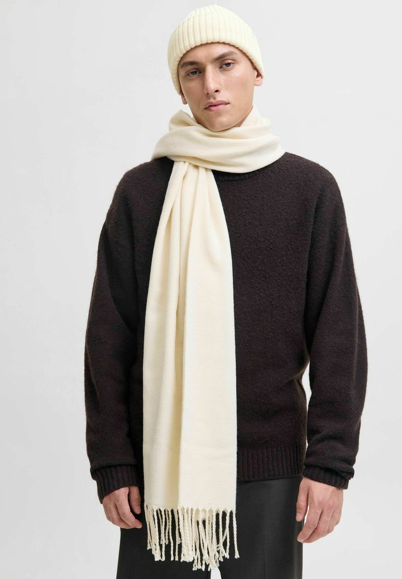 Gorro de punto acanalado beige y bufanda crema con extremos con flecos, llevados sobre un suéter texturizado marrón oscuro, contra un fondo claro y liso.