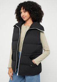 Schwarze Puffer-Weste mit Reißverschluss vorne, hohem Kragen und zwei Fronttaschen; hat eine glatte, gepolsterte Textur und weiße Reißverschlusdetails.