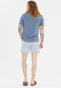 Mäns blå bomullströja, randiga badshorts och sandaler. Enastående tatueringar på armar och ben. Avslappnad, luftig passform.