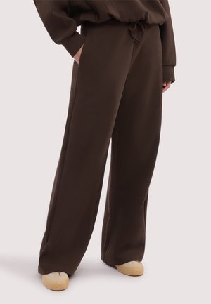 Persona che indossa pantaloni larghi color marrone scuro con le mani nelle tasche e scarpe beige con dettagli in rete, in piedi su uno sfondo chiaro.