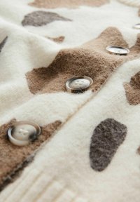 Fleece kangashuppu, jossa on kermaisa pohja ja ruskea lehmäkuvio; etuosassa on suuret, pyöreät, vaaleat napit kiinnitystä varten.