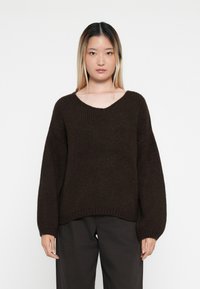 Pletený hnědý svetr s výstřihem do V, oversized rukávy a žebrovanou texturou, v kombinaci s tmavými kalhotami. Jednoduchý design bez viditelných vzorů.