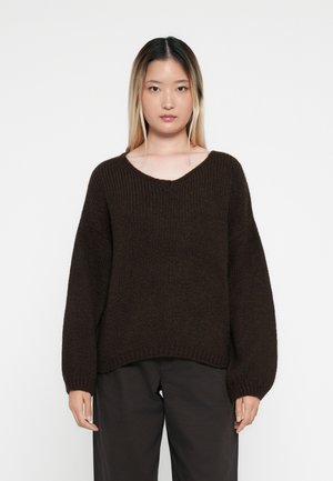 Maglione marrone lavorato a maglia con scollatura a V, maniche oversize e texture a coste, abbinato a pantaloni scuri. Design semplice senza motivi visibili.