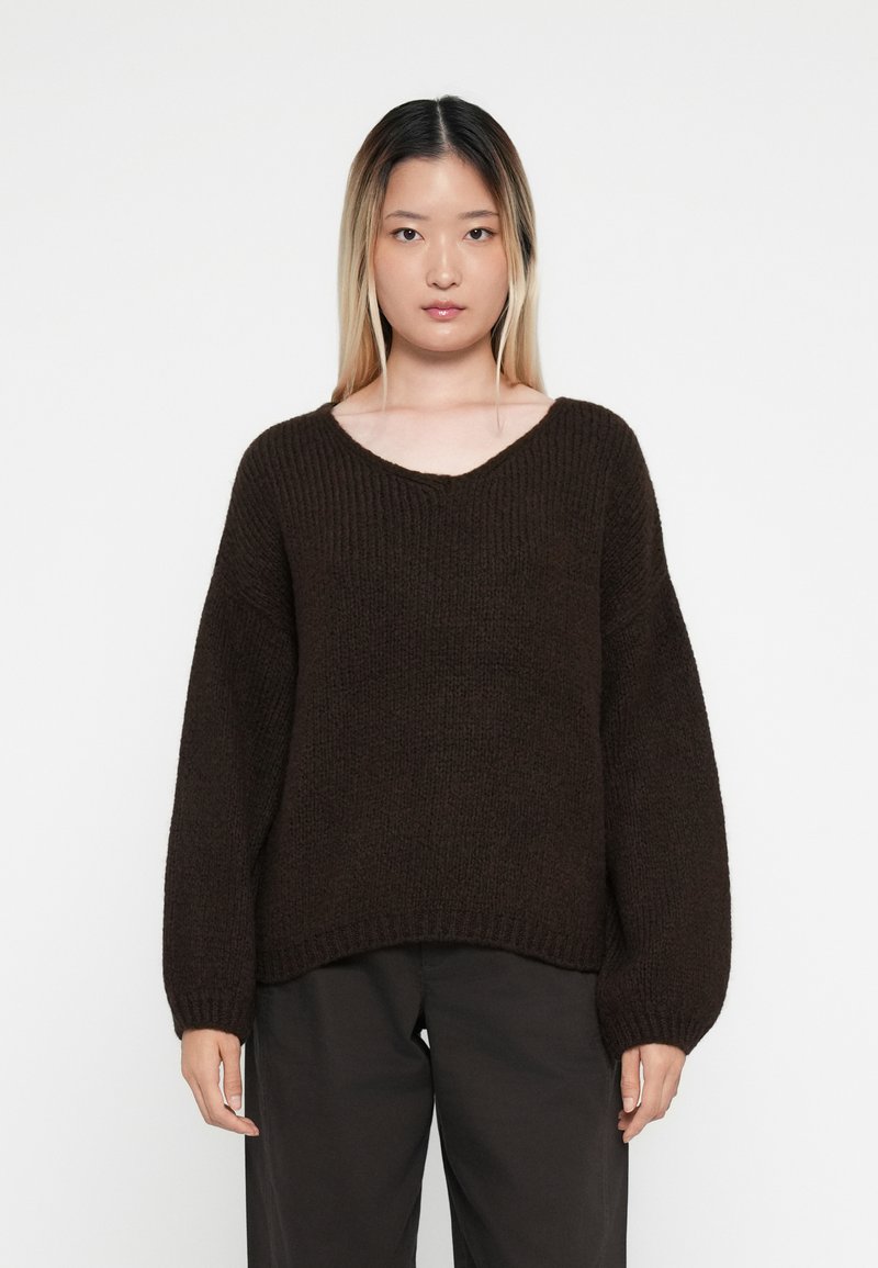 Pletený hnědý svetr s výstřihem do V, oversized rukávy a žebrovanou texturou, v kombinaci s tmavými kalhotami. Jednoduchý design bez viditelných vzorů.