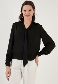 Black, long-sleeve blouse with a tie-neck detail, soft fabric, and gathered cuffs, paired with light-colored pants.