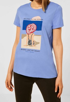 Kobieta ubrana w jasnoniebieską koszulkę z ilustracją osoby w kostiumie kąpielowym w groszki i kapeluszu z szerokim rondem siedzącej na plaży.