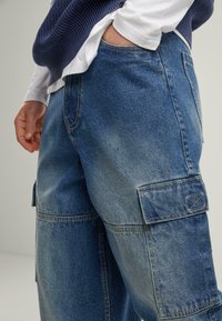 Denim cargo pants in light blue, with large side pockets and visible stitching details. Features a relaxed fit and soft texture.