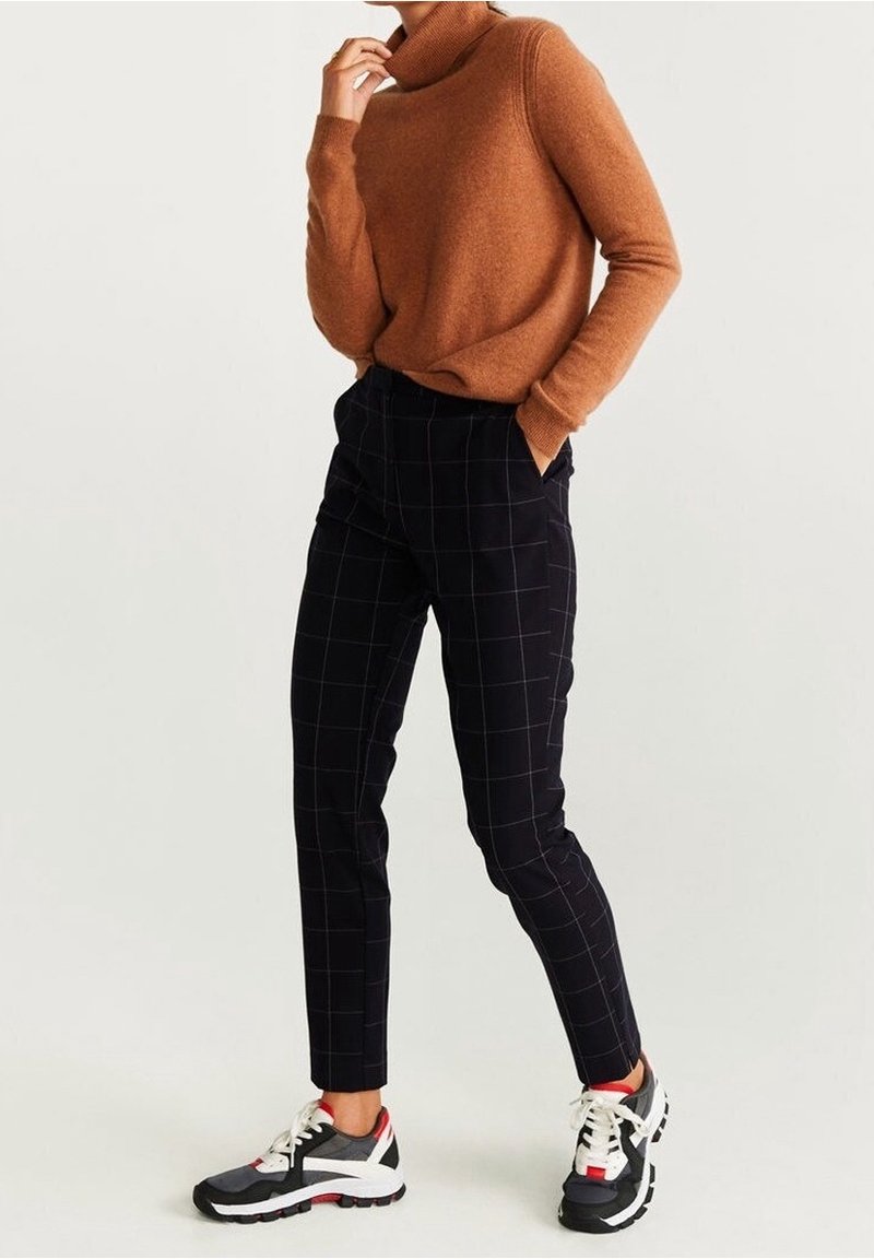Personne portant un pull à col roulé marron, un pantalon à carreaux noirs, et des baskets noires, blanches et rouges, debout avec une main dans la poche.