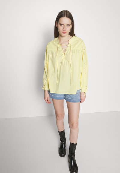 Femme portant un haut long à manches longues de couleur jaune pâle avec un décolleté lacé et des manches, un short en denim et des bottes noires mi-mollet, se tenant sur un sol gris.