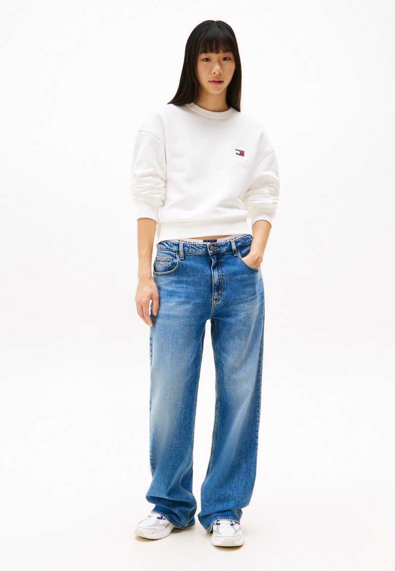 Weißer Sweatshirt mit gerippten Bündchen, blaue weit geschnittene Jeans mit verblasster Textur und weiße Sneakers mit blauen Akzenten. Das Modell steht vor einem schlichten Hintergrund.