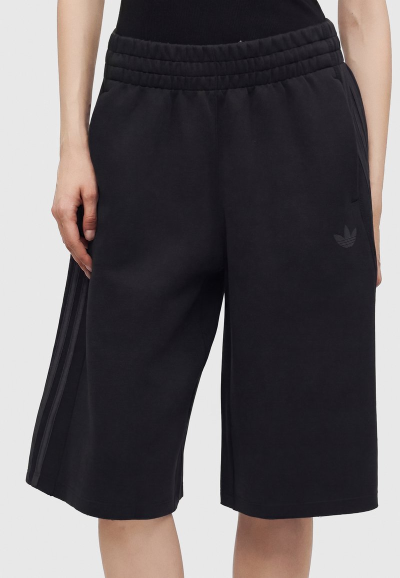 Personne portant un short noir large jusqu'aux genoux avec une taille élastique et de fines rayures latérales sur un fond uni.