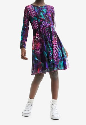 Langärmliges Kleid mit lila Blumen- und Tigerdruck auf einem strukturierten Stoff, ausgestattet mit einem ausgestellten Rock. Kombiniert mit weißen Sneakers.